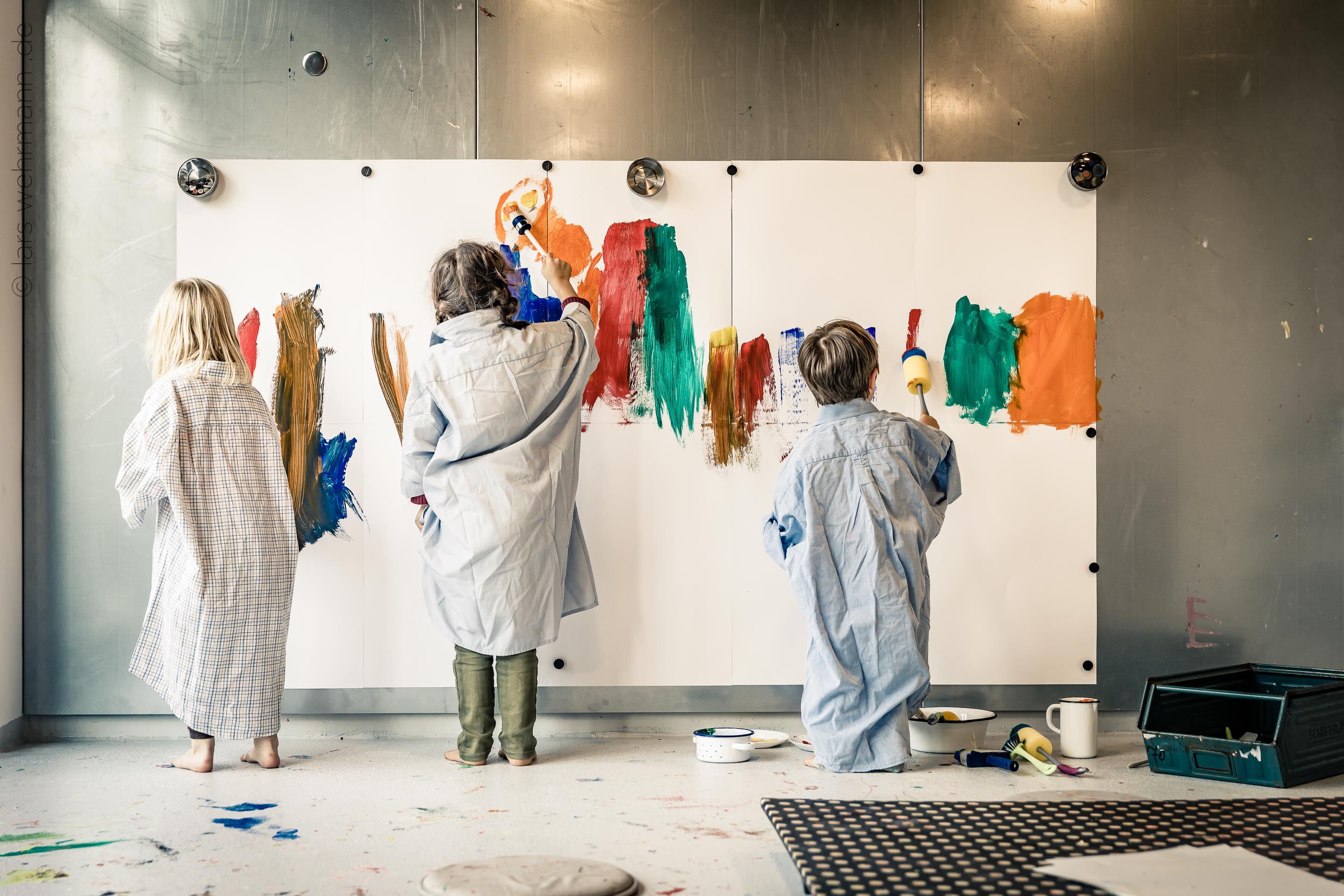 Drei Kinder in Malerkitteln malen farbenfrohe Striche auf eine große weiße Leinwand. Der Boden und die Wände sind mit Farbe bespritzt. Kreative Atmosphäre.